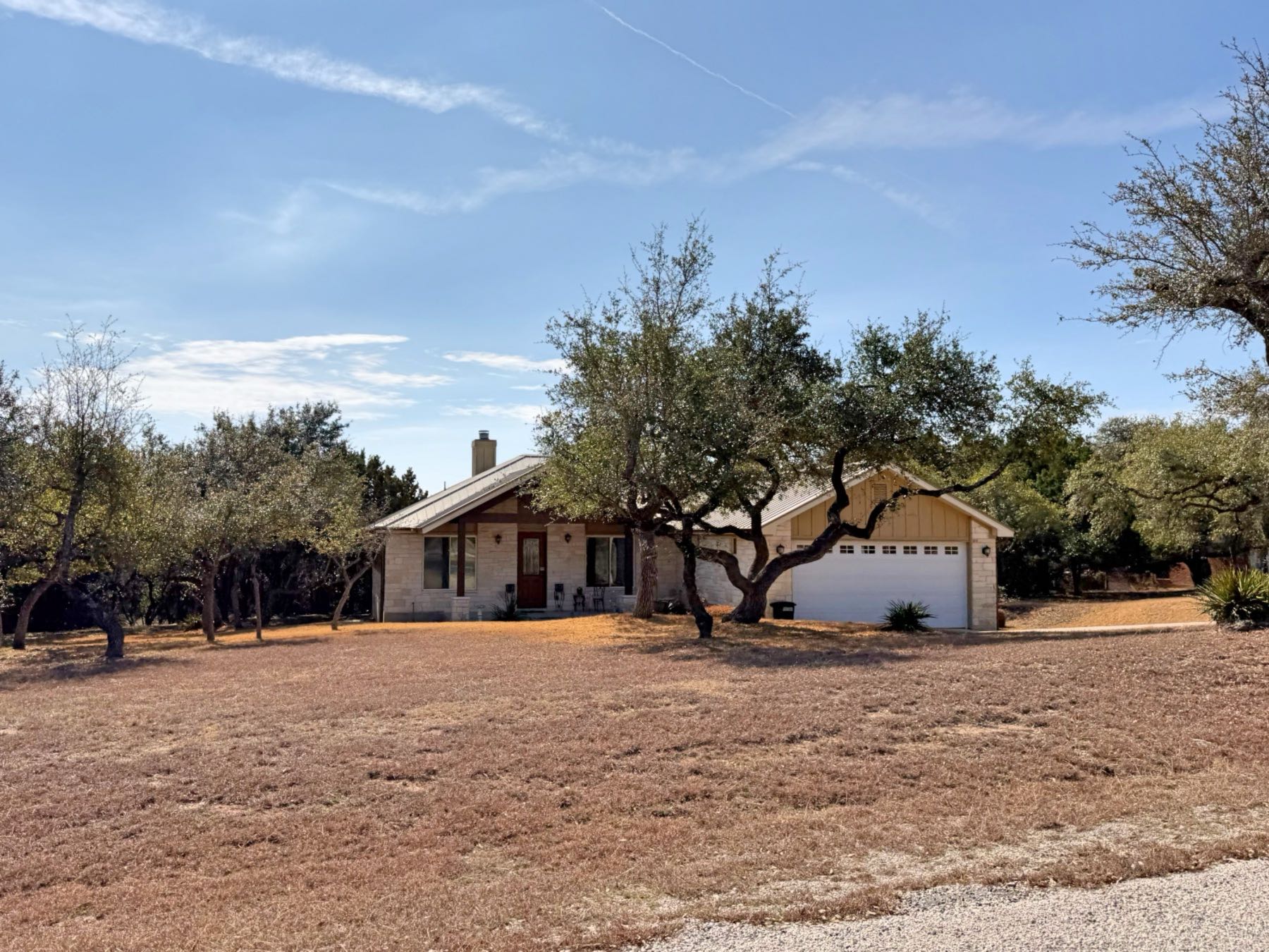 Ranch-style home with metal roof and attached garage surrounded by live oaks on large lot in Sunset Canyon, Dripping Springs TX 78620