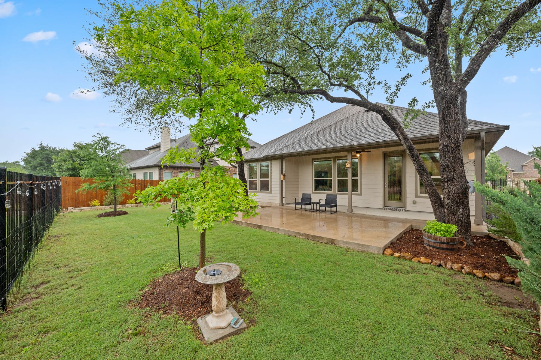 Backyard at 113 Orange Mimosa Lane Leander TX with covered concrete patio, green lawn, mature trees, iron fence, and decorative bird bath